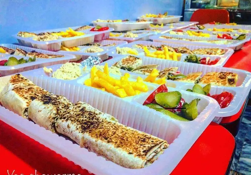 تصویری از میز پر از وعده‌های غذایی فست فود شاورما یاس شامل شاورما، سیب‌زمینی، حمص و ترشی در ظروف یکبار مصرف برای بیرون‌بر.