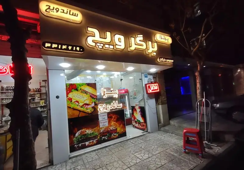 نمای شبانه فست فود برگرویچ با تابلوی روشن، عرضه کننده ساندویچ، پیتزا و فلافل، به همراه شماره تماس ۰۳۱۳۷۶۷۱۹۰ و سرویس رایگان.