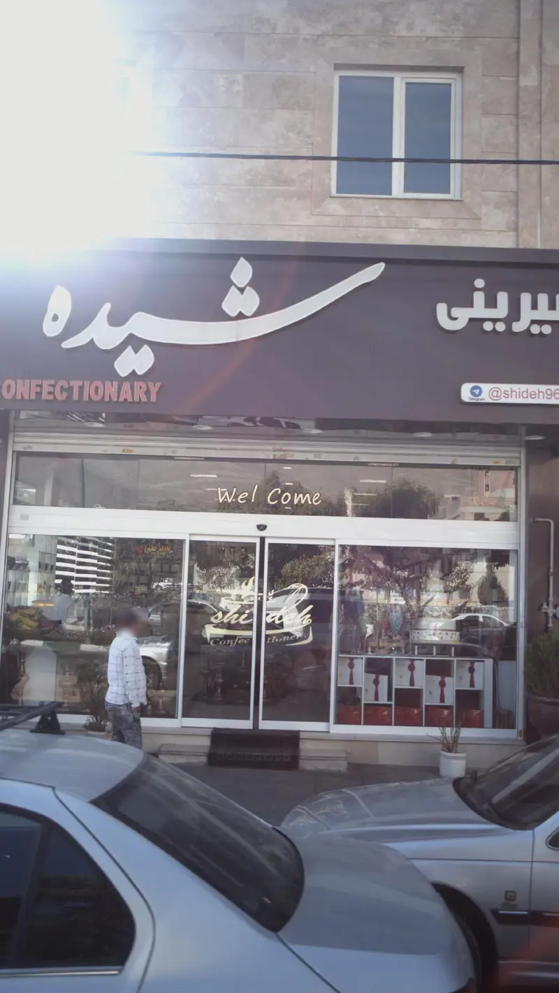 تصویری از نمای جلویی فروشگاه شیرینی شیده با تابلوی مشخص نام برند، ورودی شیشه‌ای و محصولات متنوع شیرینی‌پزی داخل ویترین.