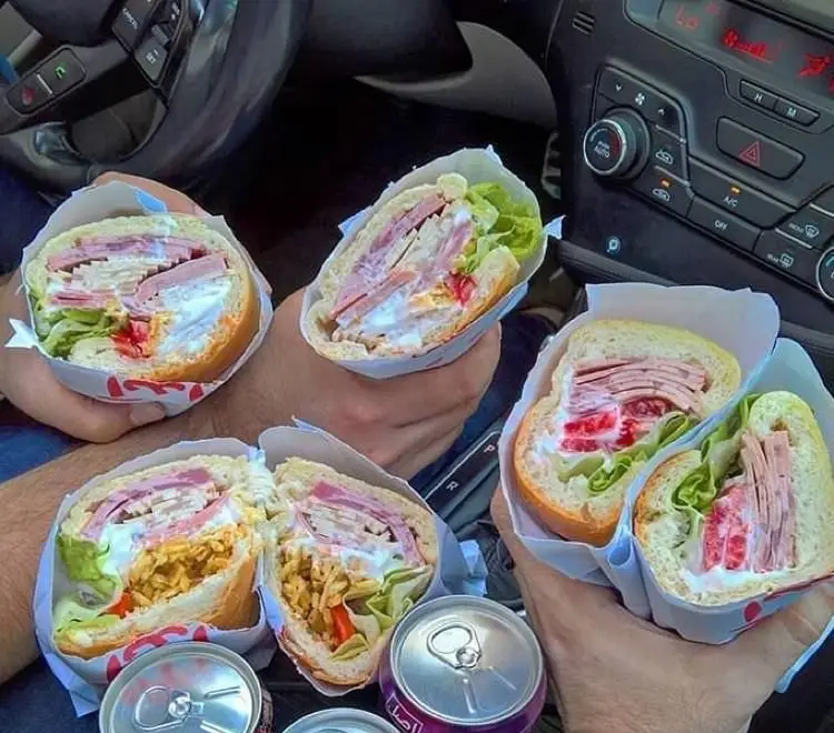 چندین ساندویچ ژامبون پر و خوشمزه ساندویچ آیدا پلاس مرزداران در دست مشتریان داخل خودرو، با نوشابه برای یک دورهمی دوستانه.