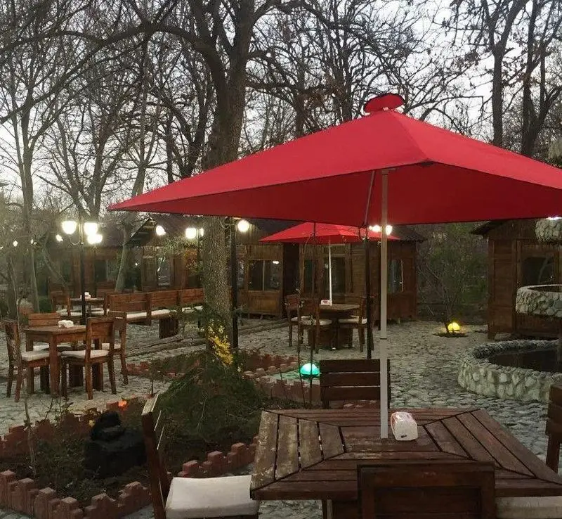 باغ رستوران شادی، فضایی زیبا و آرام با آلاچیق‌های چوبی، میز و صندلی‌های فضای باز، چترهای قرمز و آبنمای سنگی دلنشین.