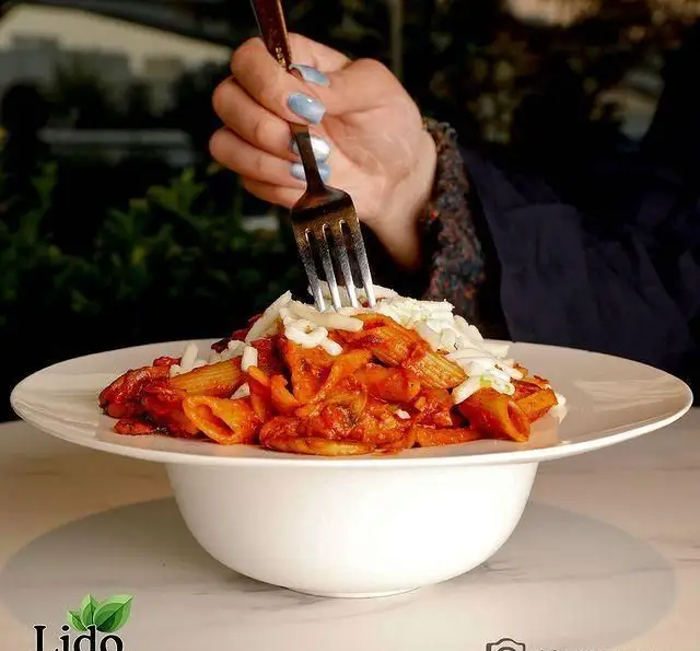 تصویری اشتهاآور از پاستا پنه با سس گوجه، پنیر رنده شده و قارچ. دست با چنگال آماده خوردن در رستوران ایتالیایی لیدو، طعمی فراموش‌نشدنی.