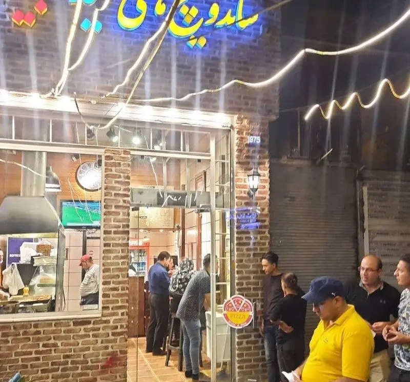 ساندویچ های حمید و مجید، فست فود محبوب با مشتریان شلوغ، تابلوی نئون درخشان و نمای آجری در شب.