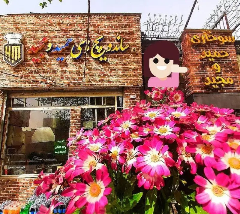 نمای بیرونی فست فود ساندویچ های حمید و مجید با ساختمان آجری، لوگو سرآشپز و گل‌های صورتی پرطراوت.