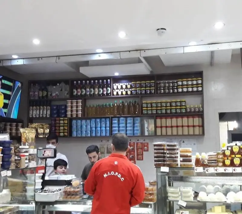 فروشگاه لبنیات سنتی دهکده: قفسه‌هایی مملو از محصولات تازه محلی، عسل، روغن، پنیر و ماست با مشتریان در حال خرید.