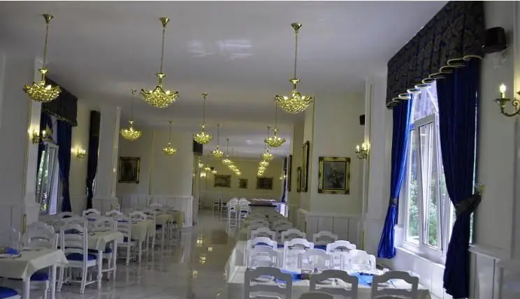 نمایی از سالن غذاخوری شیک و دلباز رستوران ایرانی استخر ونک، با چیدمان میز و صندلی‌های سفید، لوسترهای درخشان و پرده‌های آبی.