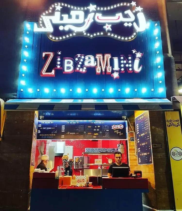 تصویری روشن و جذاب از فست فود گامبی ساندویچ، شامل تابلوی نئون زیبامینی، پیشخوان سفارشات، و منوی کامل هات داگ، سیب زمینی، برگر و نوشیدنی.