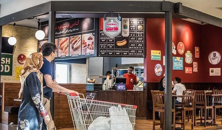 نمایی از فست فود گامبی ساندویچ با منوی متنوع شامل ساندویچ، هات داگ و برگر، همراه با مشتریان و دکوراسیون گرم و جذاب.