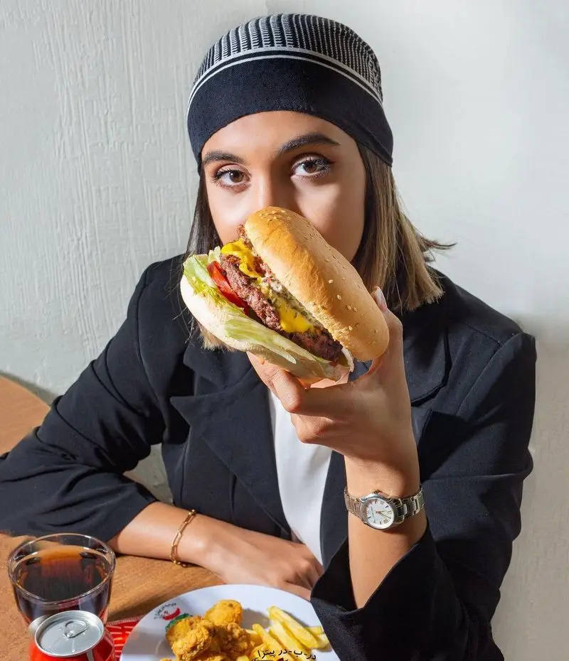 دختری جوان با هدبند، یک برگر جذاب و خوشمزه از پیتزا در ب در را در دست گرفته است. سیب زمینی، مرغ سوخاری و نوشابه در تصویر دیده می‌شود.