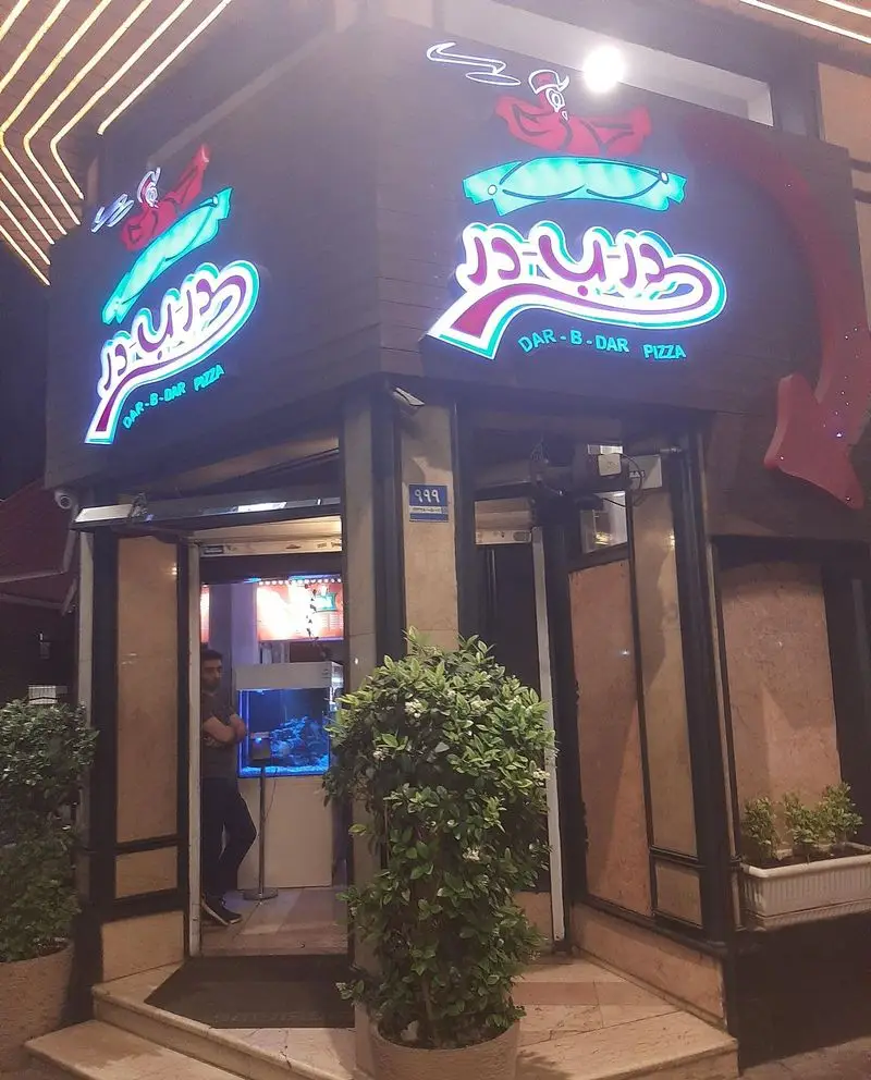 نمای بیرونی جذاب و روشن پیتزا در ب در در شب، با تابلوی نئون فارسی و انگلیسی، نشان دهنده شعبه فست فود با غذای گرم