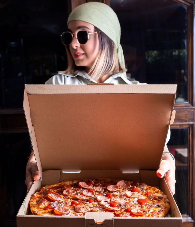 پیتزا در ب در: دختری با عینک آفتابی و روسری سبز، جعبه پیتزای خوشمزه سوسیس و پپرونی را تحویل می‌دهد. سفارش آنلاین پیتزا با بهترین کیفیت.