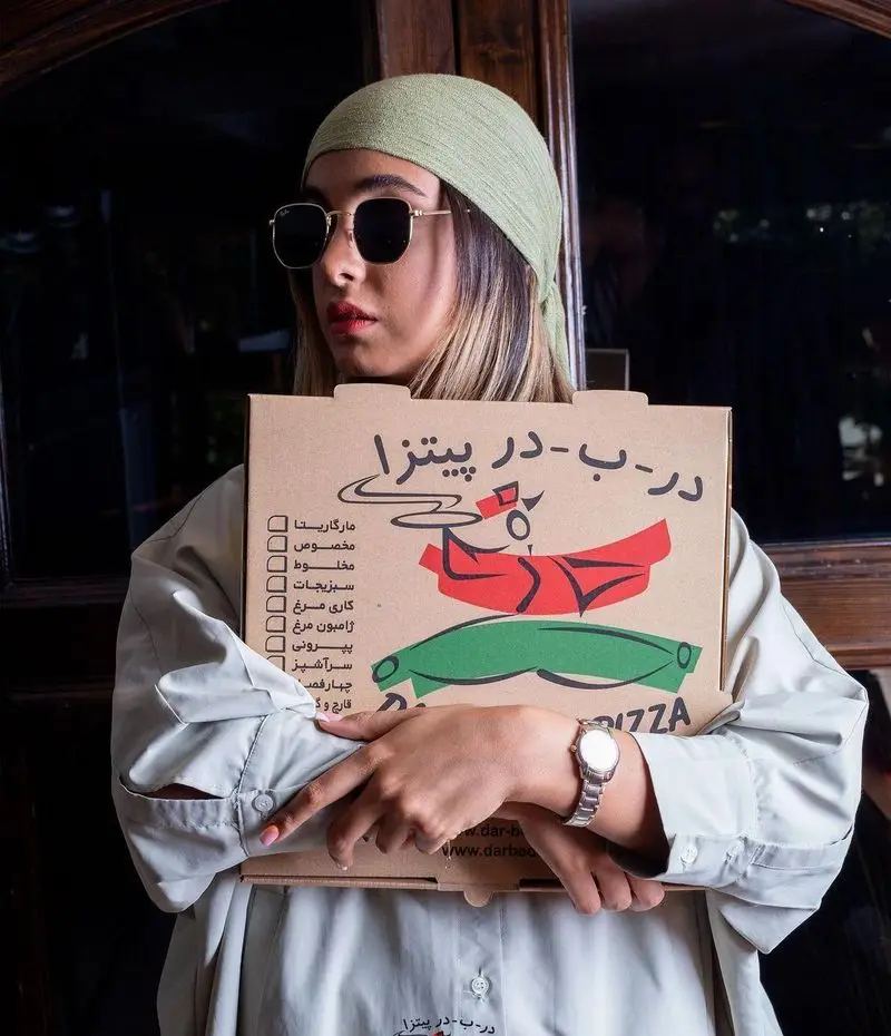 پیتزا در ب در: زن جوانی با کلاه و عینک آفتابی، جعبه پیتزای برند در ب در را در دست دارد. روی جعبه لیست انواع پیتزا و لوگو مشخص است. سفارش آنلاین پیتزا.