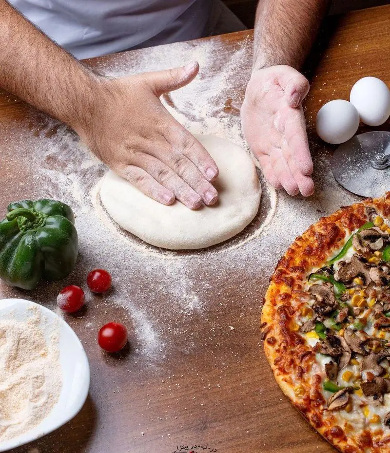 تصویری از مراحل پخت پیتزا در ب در: آماده‌سازی خمیر با دستان آشپز، همراه با پیتزای داغ قارچ و ذرت تازه. یک تجربه خوشمزه.
