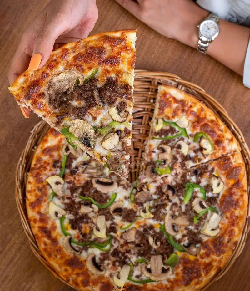 پیتزا در ب در: یک پیتزای جذاب با گوشت چرخ کرده فراوان، قارچ‌های تازه و فلفل دلمه‌ای سبز، روی خمیری طلایی و برشته. دستی برشی از آن را برمی‌دارد.