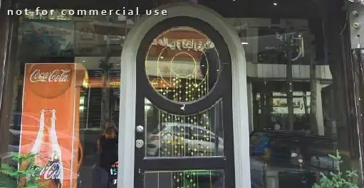 نمای بیرونی خانه پیتزا، ویترین شیشه‌ای با پوستر کوکاکولا، درب ورودی با طرح‌های دایره‌ای و نوشته عربی، بازتاب خیابان و محیط اطراف.