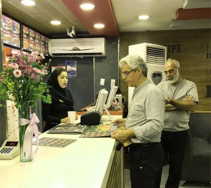 فست فود آمادو: مشتریان در حال انتخاب از منو و سفارش غذا از صندوقدار. دکوراسیون داخلی با گل و تجهیزات مدرن، محیطی دلپذیر.