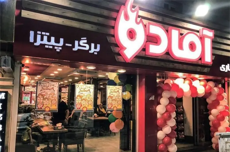 نمای بیرونی جذاب فست فود آمادو با تابلوی نئون "برگر-پیتزا"، لوگوی آتشین و ورودی تزئین شده با بادکنک‌های جشن.