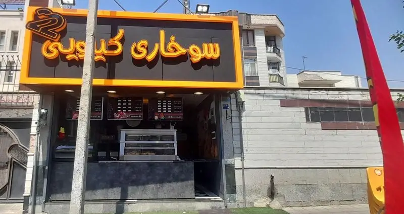 نمای بیرونی و پر جنب و جوش فروشگاه فست فود سوخاری کیلویی ۲۲ با تابلوی برجسته نارنجی، منو و ویترین آماده پذیرایی برای انواع مرغ سوخاری و غذاهای فست فودی.