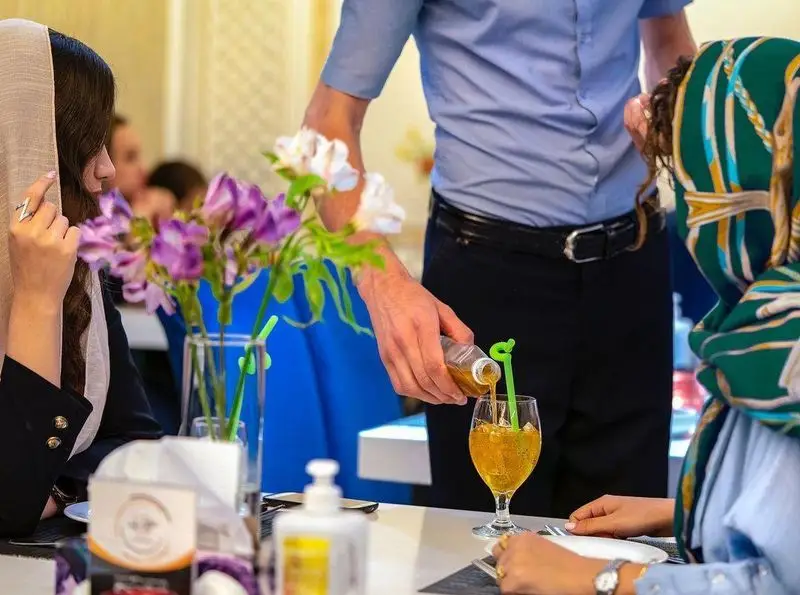 پیشخدمت در حال سرو نوشیدنی خنک در رستوران محسن، در محیطی دلنشین با گلهای تزئینی. مشتریان از خدمات عالی و فضای آرام لذت میبرند.