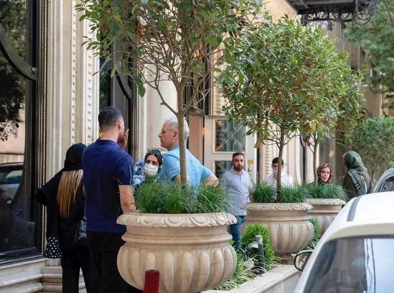مشتریان در فضای بیرونی رستوران محسن در تهران با گلدان‌های تزئینی و درختان سرسبز، محیطی دلنشین و شیک کنار ورودی رستوران.