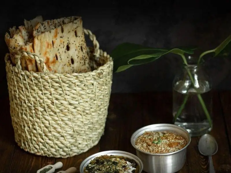 سفره‌ای سنتی با نان تازه و غذاهای ایرانی قیمه و قورمه در کنار سبد حصیری. بهترین تجربه در رستوران قیمه قورمه.