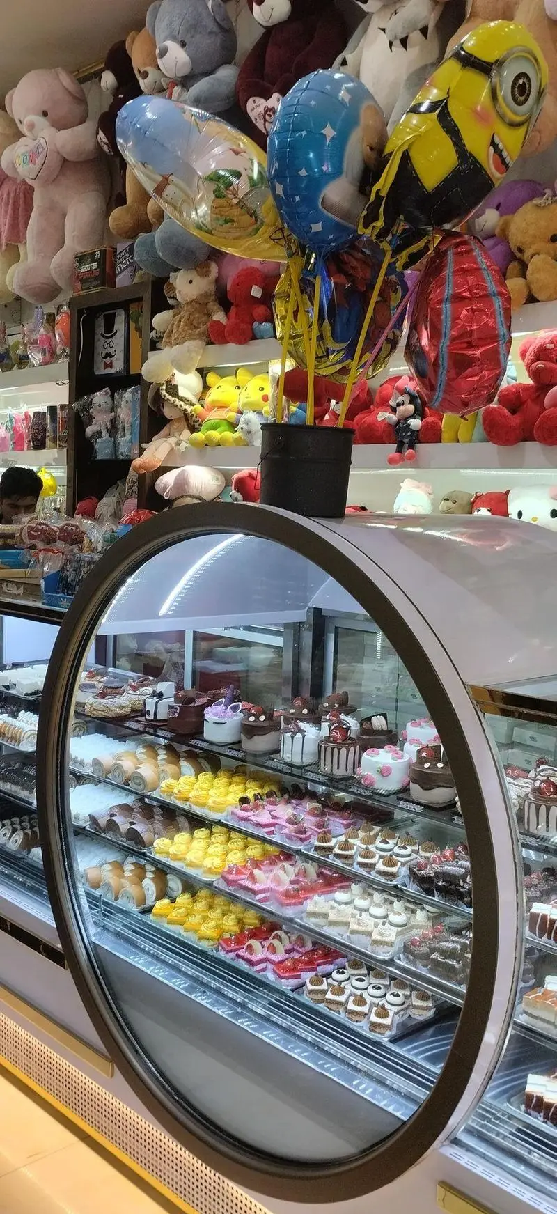 ویترین شیرینی کندو پر از کیک‌های رنگارنگ، دسر و انواع شیرینی تازه در کنار عروسک‌های جذاب و بادکنک‌های فانتزی آماده برای هدیه.