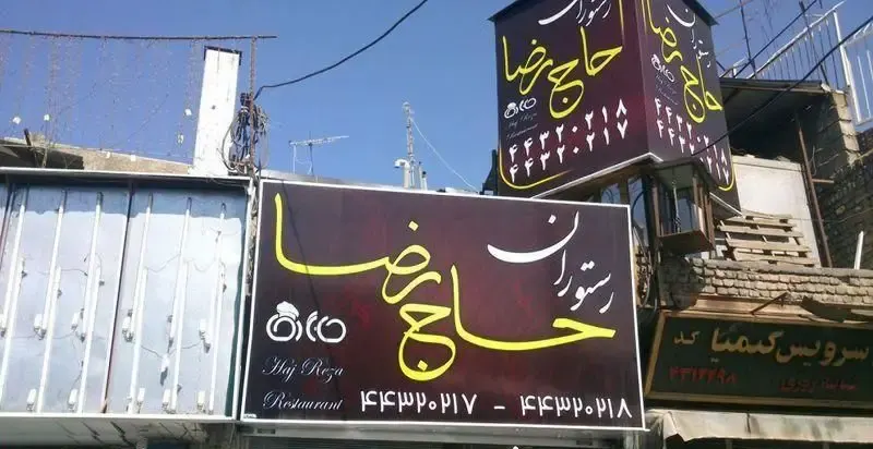 تصویر نمای بیرونی رستوران حاج رضا با تابلوهای بزرگ و روشن. این رستوران میزبان شما برای تجربه‌ی غذای سنتی و لذیذ ایرانی است.