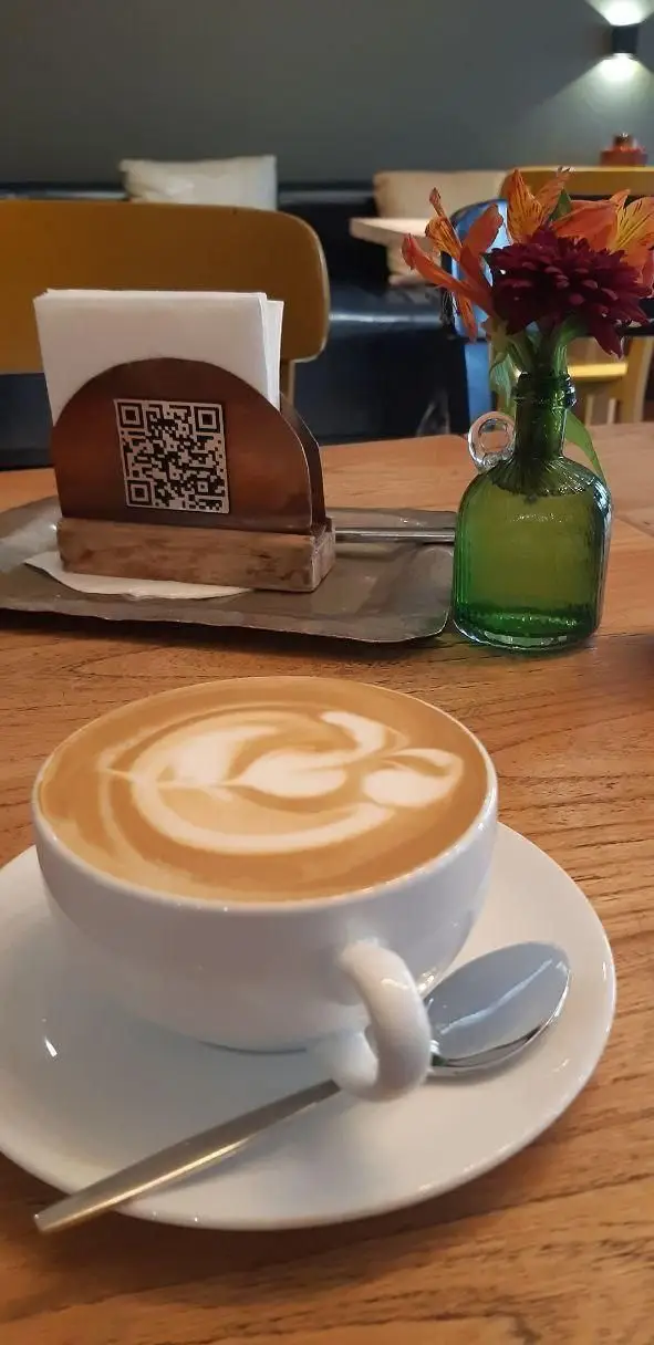 یک فنجان قهوه لاته آرت با طرح هنری در فضای دلنشین رستوران سبوس، روی میز چوبی با تزئینات زیبا و گلدان گل‌های تازه.