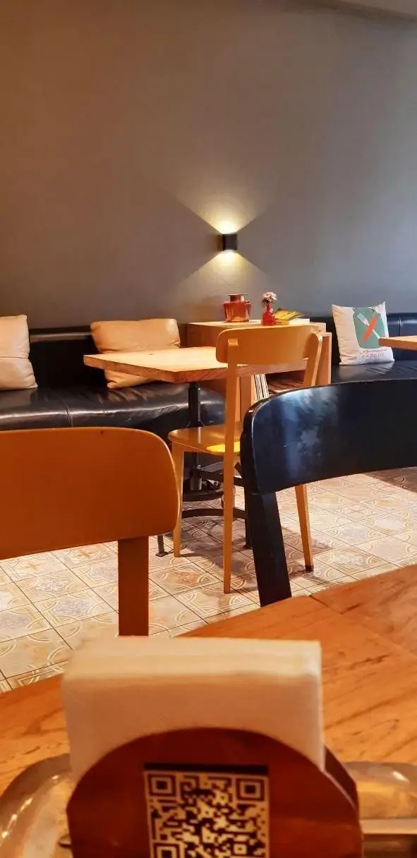 فضای داخلی رستوران سبوس با میزهای چوبی، صندلی‌ها، نیمکت چرمی و نورپردازی دیواری. کفپوش طرح‌دار و کد QR منو روی میز قابل مشاهده است.