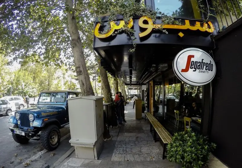 نمای بیرونی کافه و رستوران سبوس در یک روز آفتابی، با تابلوی 'سبوس' و 'Segafredo'، نیمکت چوبی، درختان و جیپ رنه‌گید آبی رنگ.