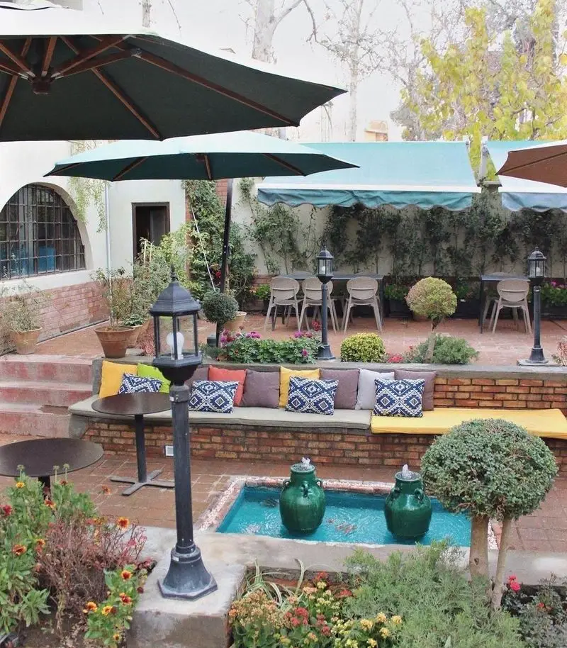 فضای روباز و زیبای کافه تی آرت با حیاطی سرسبز، چترهای بزرگ، نشیمن‌های راحت با کوسن‌های رنگی، میز و صندلی و حوض کوچک. مکانی آرامش‌بخش.