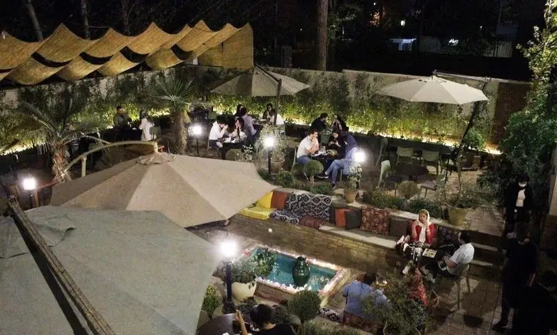 تصویری دلنشین از کافه تی آرت در شب، با مشتریان در فضای باز دنج و سرسبز، میزهای چوبی و حوض آب، مکانی ایده‌آل برای دورهمی