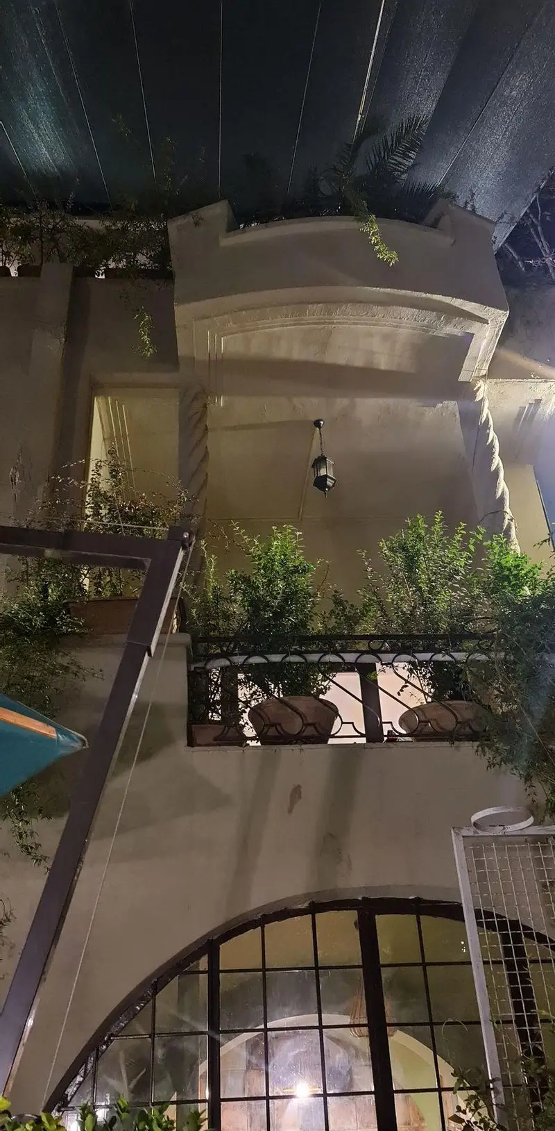 نمای شبانه کافه تی آرت با معماری خاص، شامل بالکن سرسبز، ستون های تزئینی و نورپردازی دلنشین فانوس. فضایی آرام و دنج برای مشتریان.