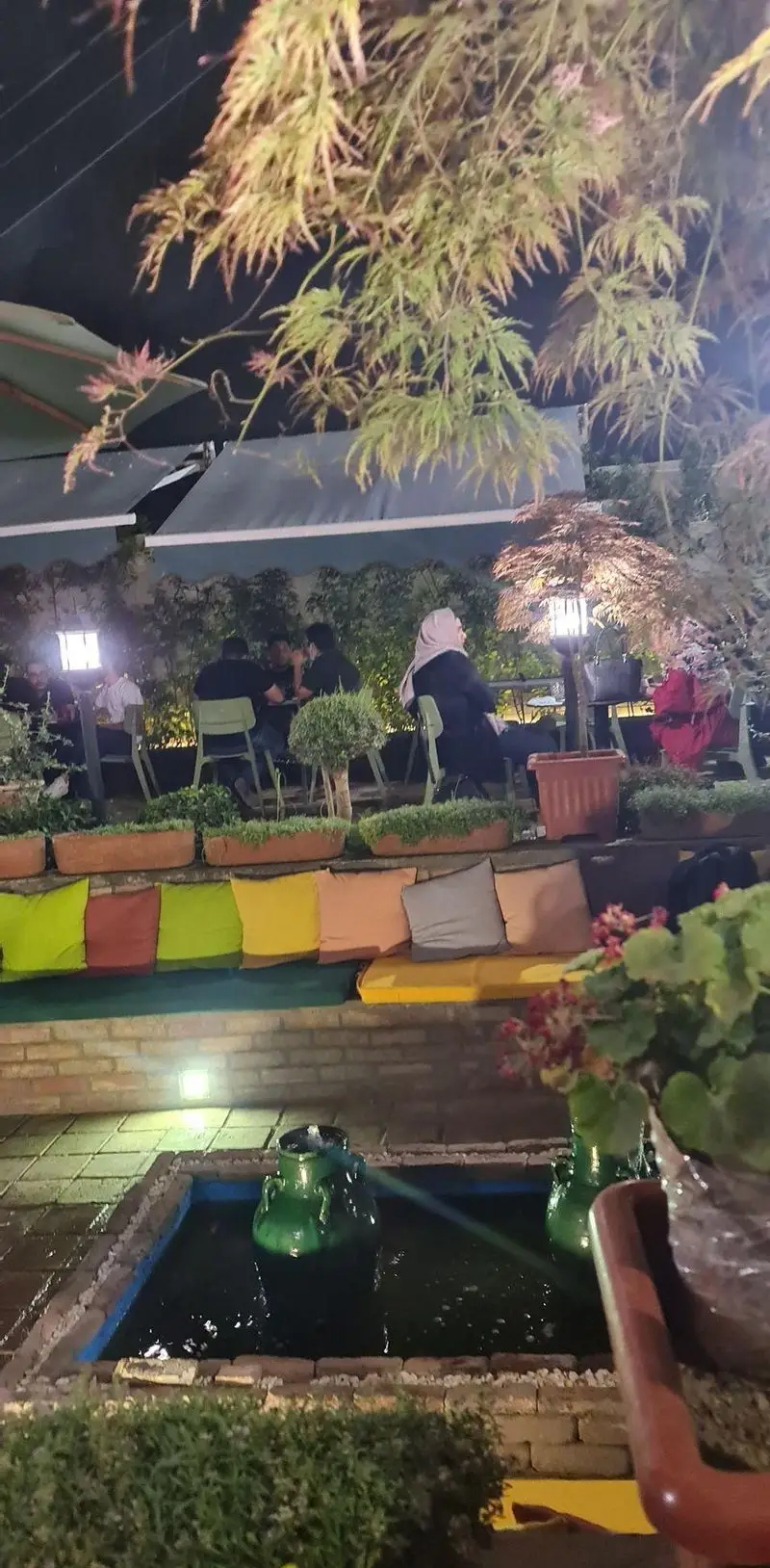 فضای دنج و زیبای کافه تی آرت در شب، میزبان دورهمی صمیمانه مشتریان در فضای باز با نورپردازی آرامش بخش و گل و گیاه.