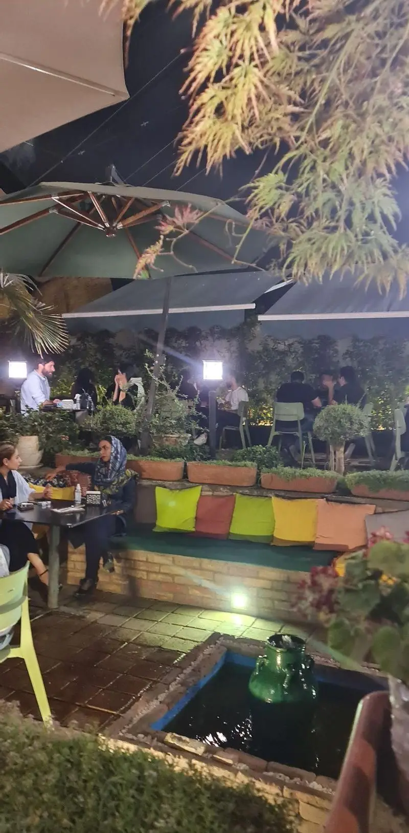 فضای دنج و سرسبز کافه تی آرت در شب با مشتریان، میز و صندلی، چترهای بزرگ، گیاهان و آبنما. تجربه‌ای دلنشین و آرامش‌بخش در فضای باز.