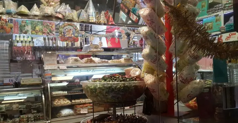 قنادی جهان نما، نمای داخلی شلوغ و جذاب با ویترین‌های پر از انواع شیرینی‌های تازه و دسر. همچنین شامل خشکبار و لوازم جشن برای مشتریان.