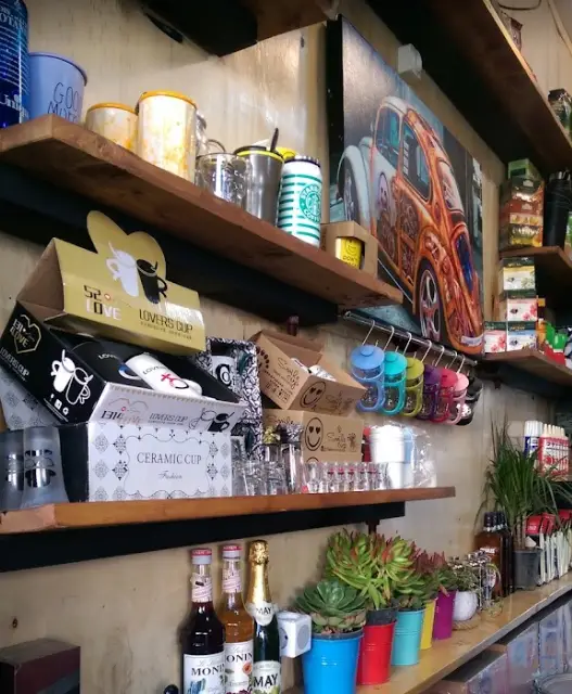 قفسه های شلوغ و جذاب کافه امیر: لیوان های طرح دار، ماگ قهوه، سیروپ و نوشیدنی، گل و گیاه و تابلوی ماشین کلاسیک فولکس واگن.