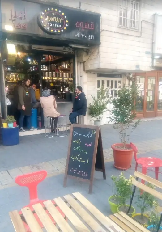 نمای کافه امیر (Amir Cafe) در پیاده‌رو با تابلوی Amuy Coffee، مشتریان در حال لذت بردن از قهوه، میز و صندلی چوبی و گلدان‌های زینتی فضای باز.