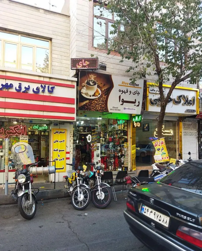 تصویری از نمای بیرونی کافه ریوا (چای و قهوه) در یک خیابان ایرانی با تابلو و ویترین جذاب، موتورسیکلت و خودروی پارک شده.