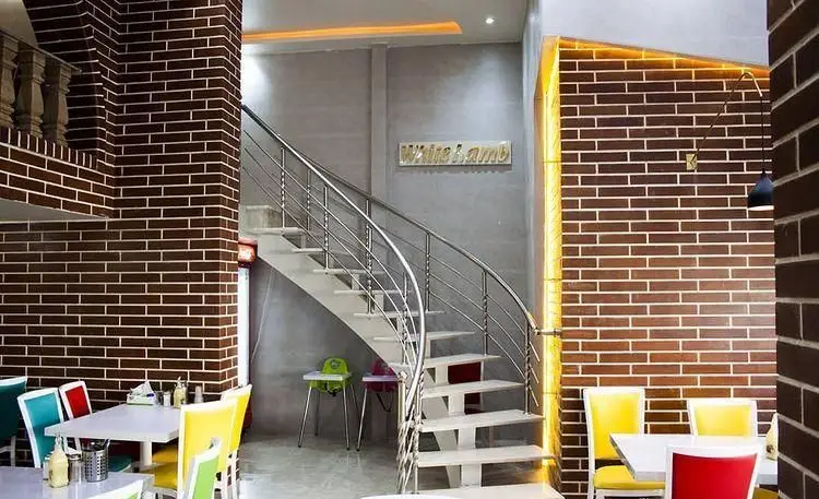 محیط داخلی طباخی بره سفید با دکوراسیون مدرن شامل دیوار آجری، پله‌های منحنی نقره‌ای، میز و صندلی‌های رنگی و تابلوی White Lamb.