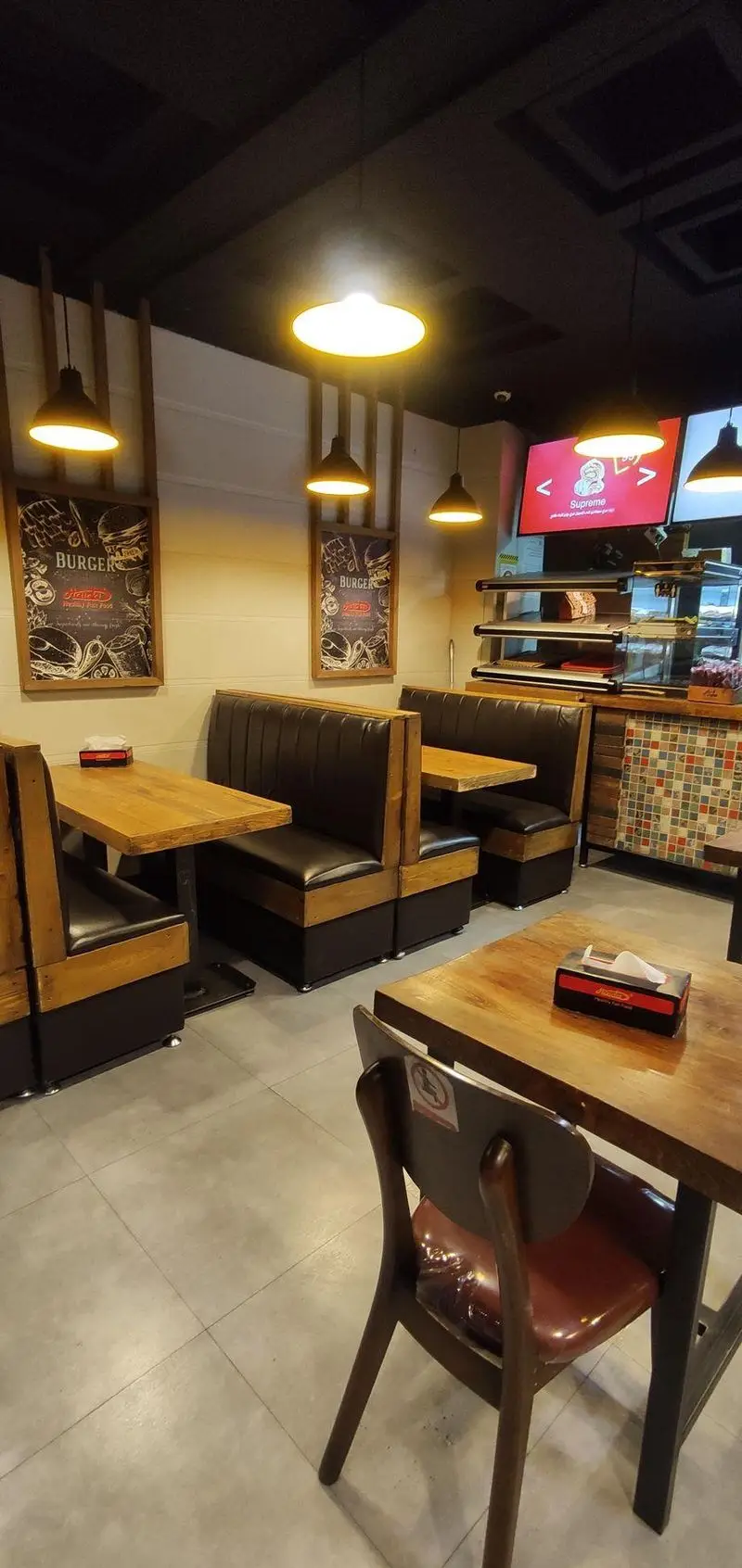 دکوراسیون شیک و مدرن فست فود ساندویچ هایدا: میزهای چوبی، نیمکت‌های چرم مشکی، نورپردازی زیبا و پوسترهای جذاب برگر.