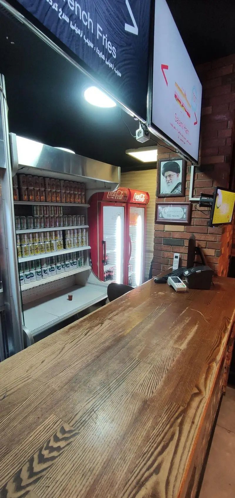 تصویر فضای داخلی ساندویچ هایدا شامل کانتر فروش چوبی، منوی دیجیتال فست فود و یخچال‌های پر از نوشیدنی آماده.