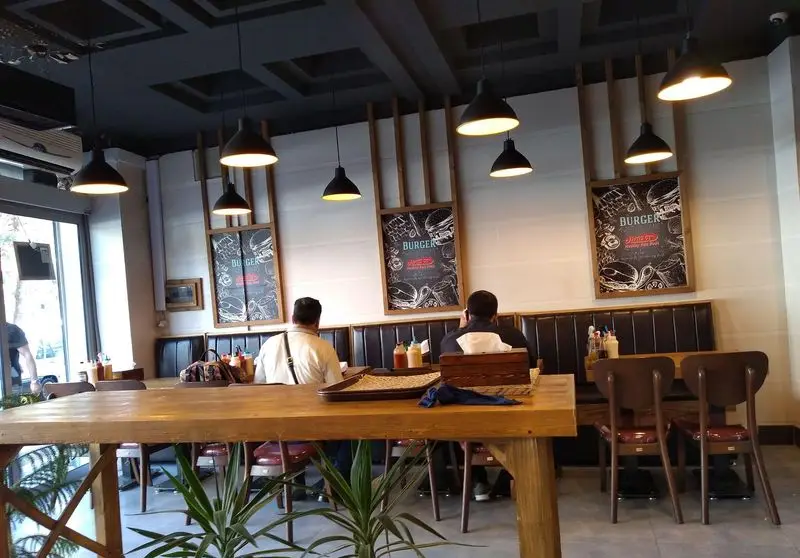 فضای داخلی دنج رستوران ساندویچ هایدا با دکوراسیون مدرن، میزهای چوبی بزرگ، نیمکت‌های تیره و تابلوهای منوی برگر.