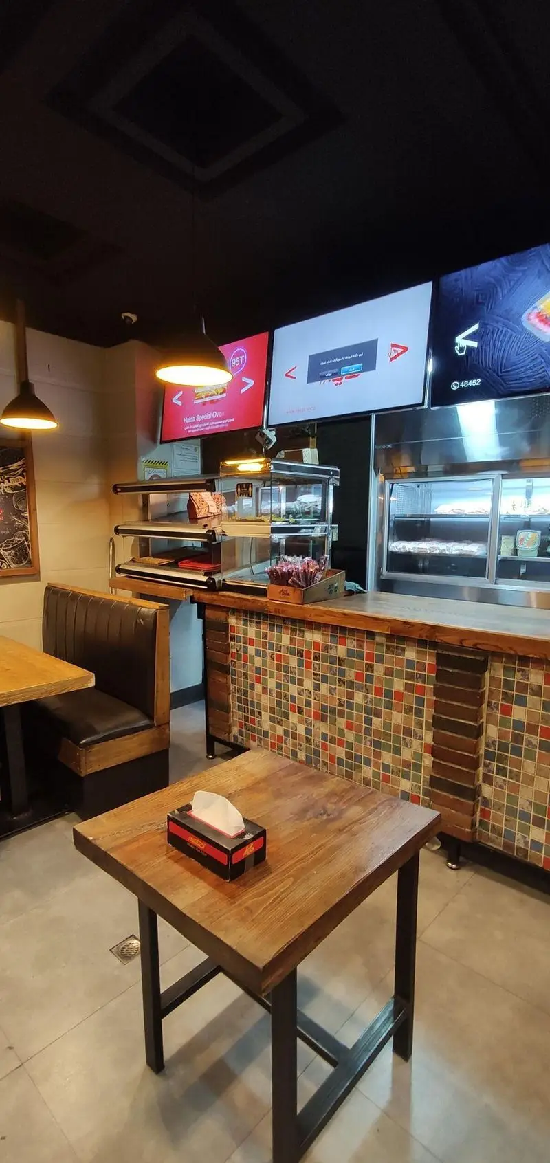 فضای داخلی دنج رستوران ساندویچ هایدا با میزهای چوبی مدرن، صندلی‌های راحتی، منوهای دیجیتال و پیشخوان سرو غذا با طراحی کاشی‌کاری جذاب.