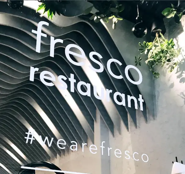 تابلوی شیشه‌ای رستوران فرسکو با نوشته "fresco restaurant" و هشتگ "#wearefresco". دکوراسیون داخلی مدرن با پنل‌های موج‌دار و گیاهان آویزان دیده می‌شود.