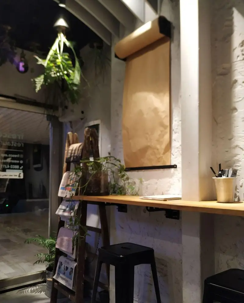 تصویری از فضای داخلی رستوران و کافه فرسکو، شامل میز بار چوبی بلند، صندلی‌های مدرن، رول کاغذ دیواری قهوه‌ای و قفسه روزنامه.