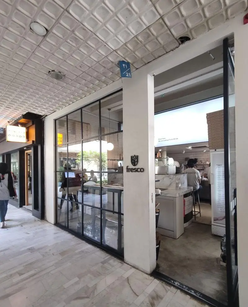 تصویر ورودی رستوران مدرن فرسکو با نمای شیشه‌ای بزرگ، لوگوی Fresco روی ستون سفید و مشتریانی در حال لذت بردن از فضای داخلی.