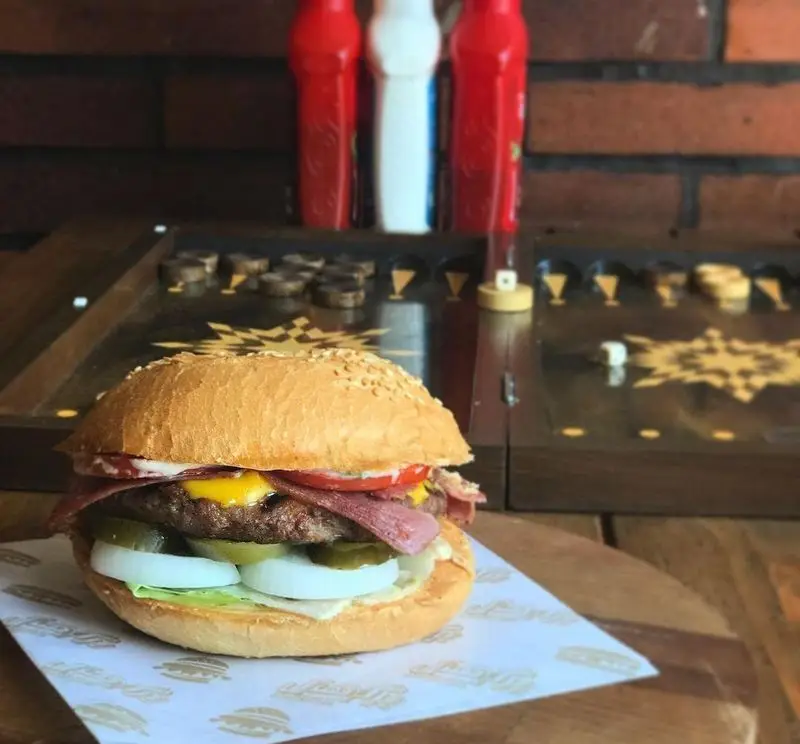 تصویری از یک همبرگر لذیذ و پرملات از فست فود برگر هال با نان کنجدی، پنیر ذوب‌شده، گوجه، خیارشور و پیاز. لذت طعم واقعی را با برگر هال تجربه کنید.