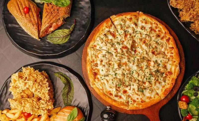 میز چیدمان شده با انواع غذاهای فست فود د ل پ: پیتزای خوشمزه مرغ و قارچ، ساندویچ گریل و مرغ سوخاری با سیب‌زمینی و سالاد. مناسب برای سفارش آنلاین.