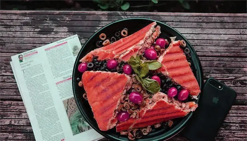 ساندویچ گریل خوشمزه فست فود د ل پ روی میز چوبی با زیتون و تزئینات، مناسب برای یک وعده غذایی لذیذ کنار روزنامه و موبایل.